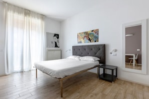 Une chambre moderne avec un lit, une petite table de chevet, un radiateur, une œuvre d’art accrochée au mur et un miroir qui reflète un bureau et une chaise.