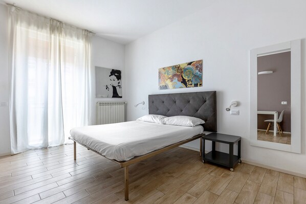 Une chambre moderne avec un lit, une petite table de chevet, un radiateur, une œuvre d’art accrochée au mur et un miroir qui reflète un bureau et une chaise.