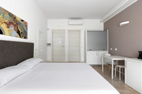 A modern hotel room with a large bed, a desk with a chair, a window with blinds, and a colorful artwork on the wall.