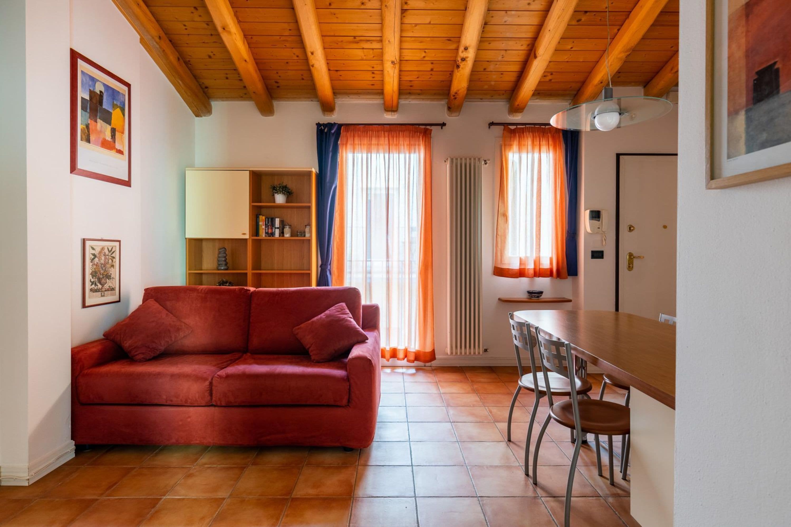 A cozy living room with a red sofa, wooden ceiling beams, a dining table with chairs, and a bookshelf.