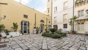 Un patio con pavimento de piedra, bancos, plantas en macetas y un edificio con fachada amarilla.