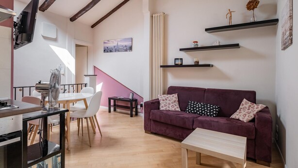Una sala de estar con un sofá morado, una mesa de centro de madera, una zona de comedor con una mesa y sillas blancas, y una fotografía de un paisaje urbano en la pared.