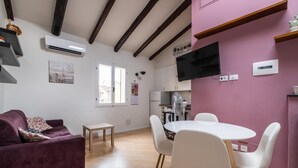 Una sala de estar moderna con un sofá morado, sillas blancas, una mesa de comedor de madera y una zona de cocina con televisor y equipo de aire acondicionado.