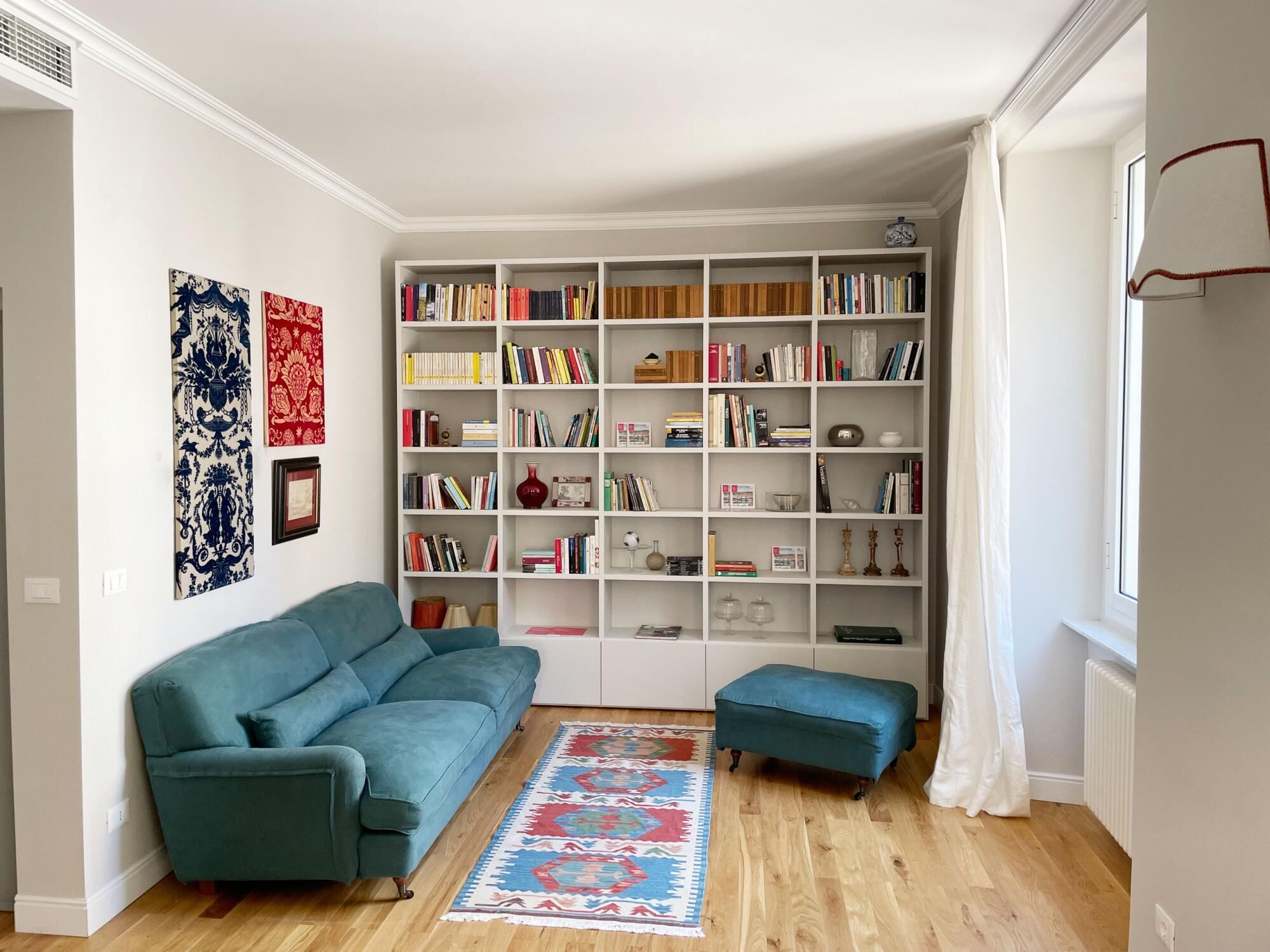 A living room with a teal sofa, a matching ottoman, a bookshelf filled with books, and a patterned rug.