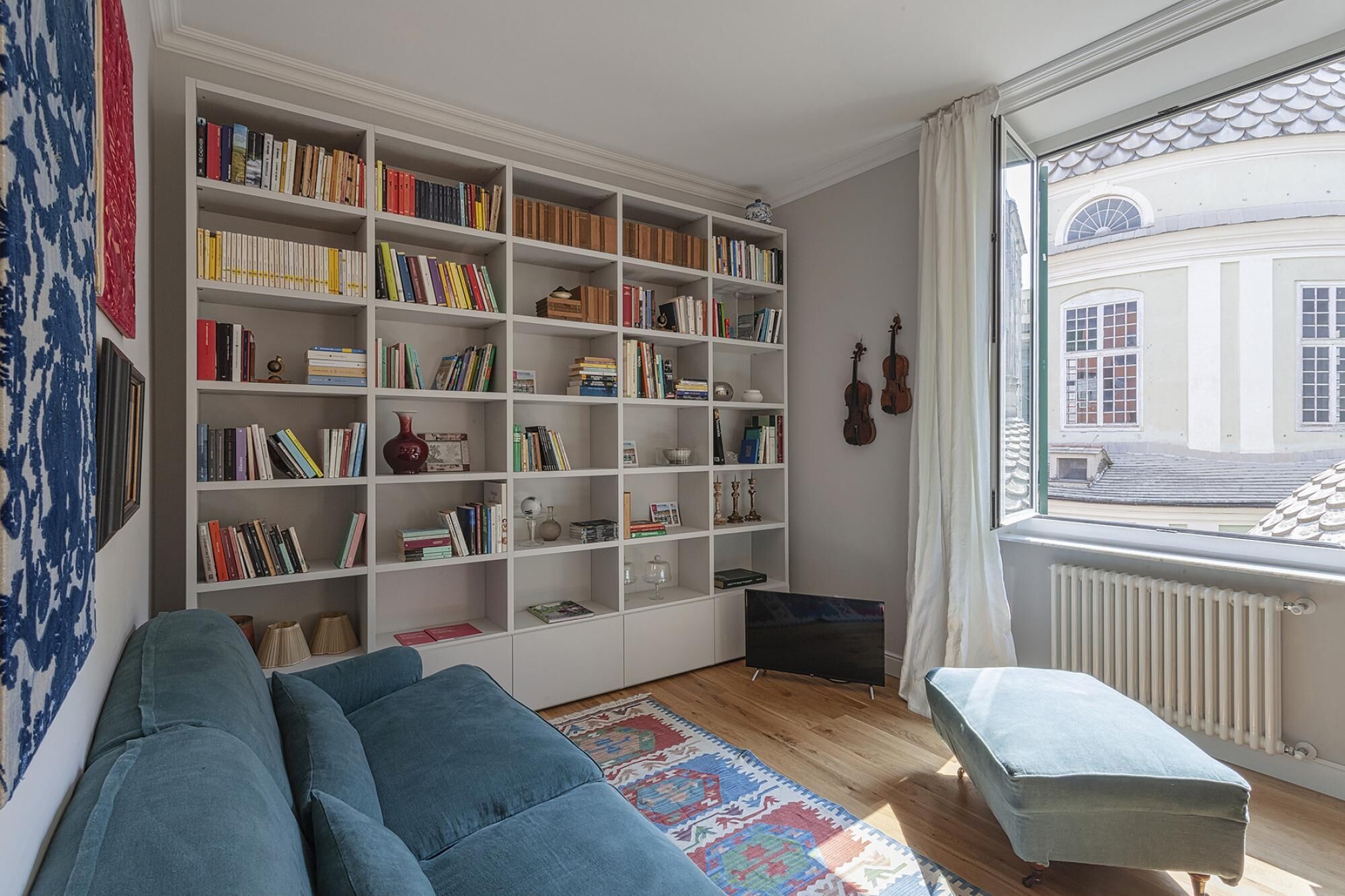 A cozy living room with a large bookshelf, a blue sofa, and a window with a view of a building.