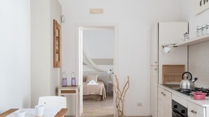 Eine kleine Küche mit weißen Schränken, einem Holztisch und einem Bett im angrenzenden Zimmer.