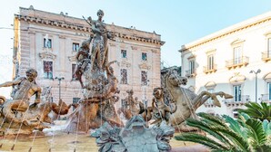 Ein klassischer Brunnen mit mehreren Statuen, umgeben von Gebäuden im klassischen Stil.
