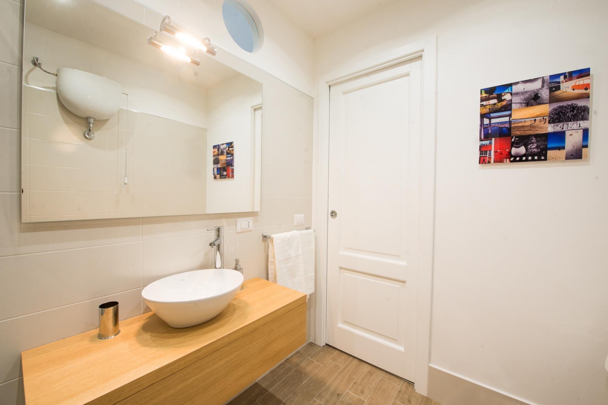 A bathroom with a white sink, wooden vanity, and a large mirror. There is a door with a poster on the wall.