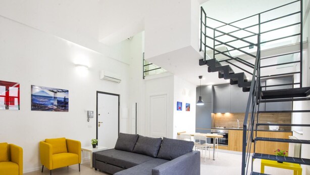 A modern apartment with a living area featuring a sofa and two chairs, a staircase, and a kitchen area.