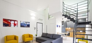Un apartament modern amb zona de saló que inclou un sofà i dues cadiretes, una escala i una cuina.