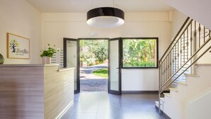 A hotel reception area with a wooden reception desk, a potted plant, and a view of the garden through glass doors. - L Ulivo Blu - One-bedroom With Balcony by Wonderful Italy (Castellammare del Golfo)