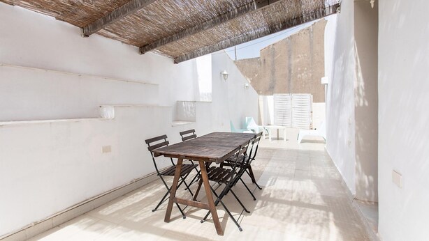 A covered outdoor patio with a wooden table and chairs, white walls, and a thatched roof.