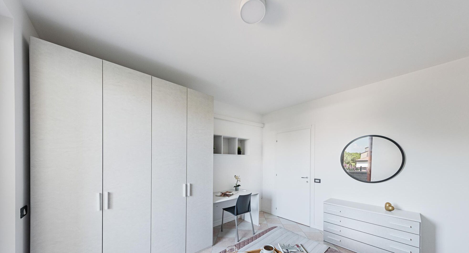 A modern bedroom with a large wardrobe, a desk with a chair, a dresser, and a round mirror on the wall.
