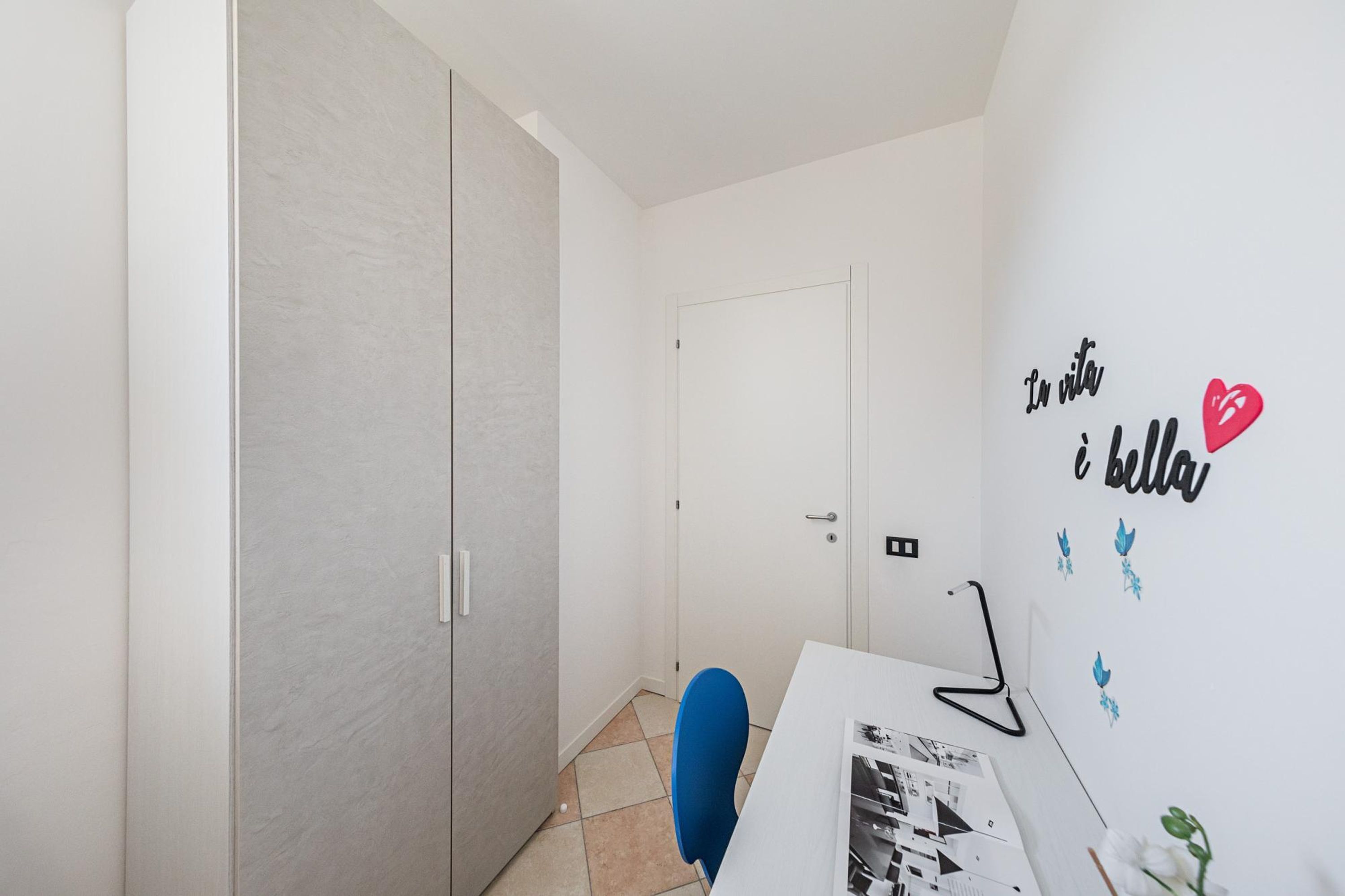 Une petite pièce moderne avec un bureau blanc, une chaise bleue et une armoire. Un élément décoratif mural présente le texte « In vita e bella » et un symbole en forme de cœur.