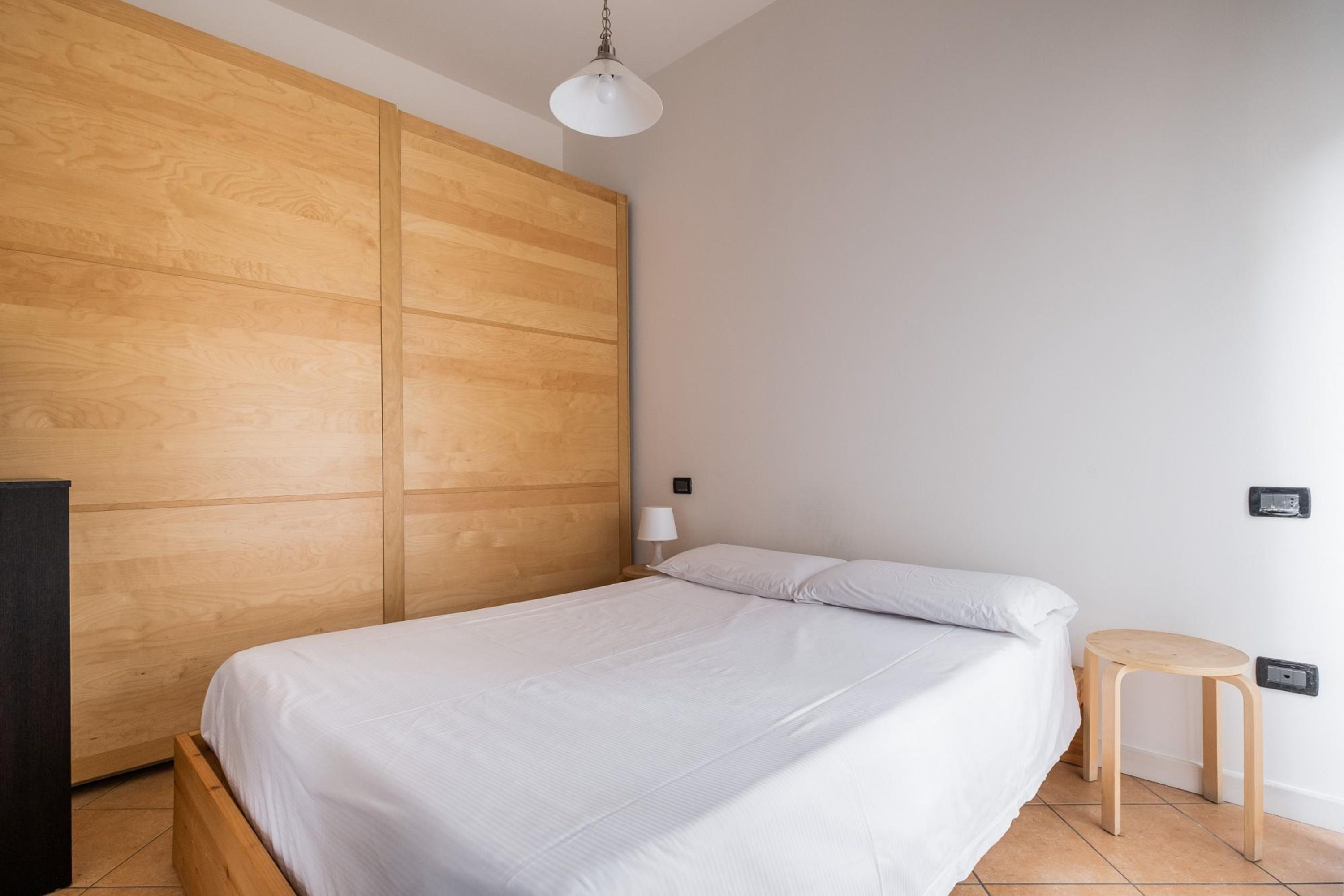 A bedroom with a wooden wardrobe, a bed with white bedding, a small wooden stool, and a white lamp.