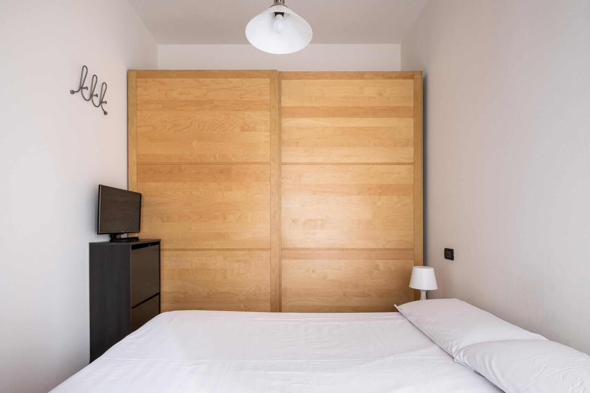 Un dormitorio con un armario de madera, un televisor sobre un mueble, una cama con ropa de cama blanca y una lámpara blanca.