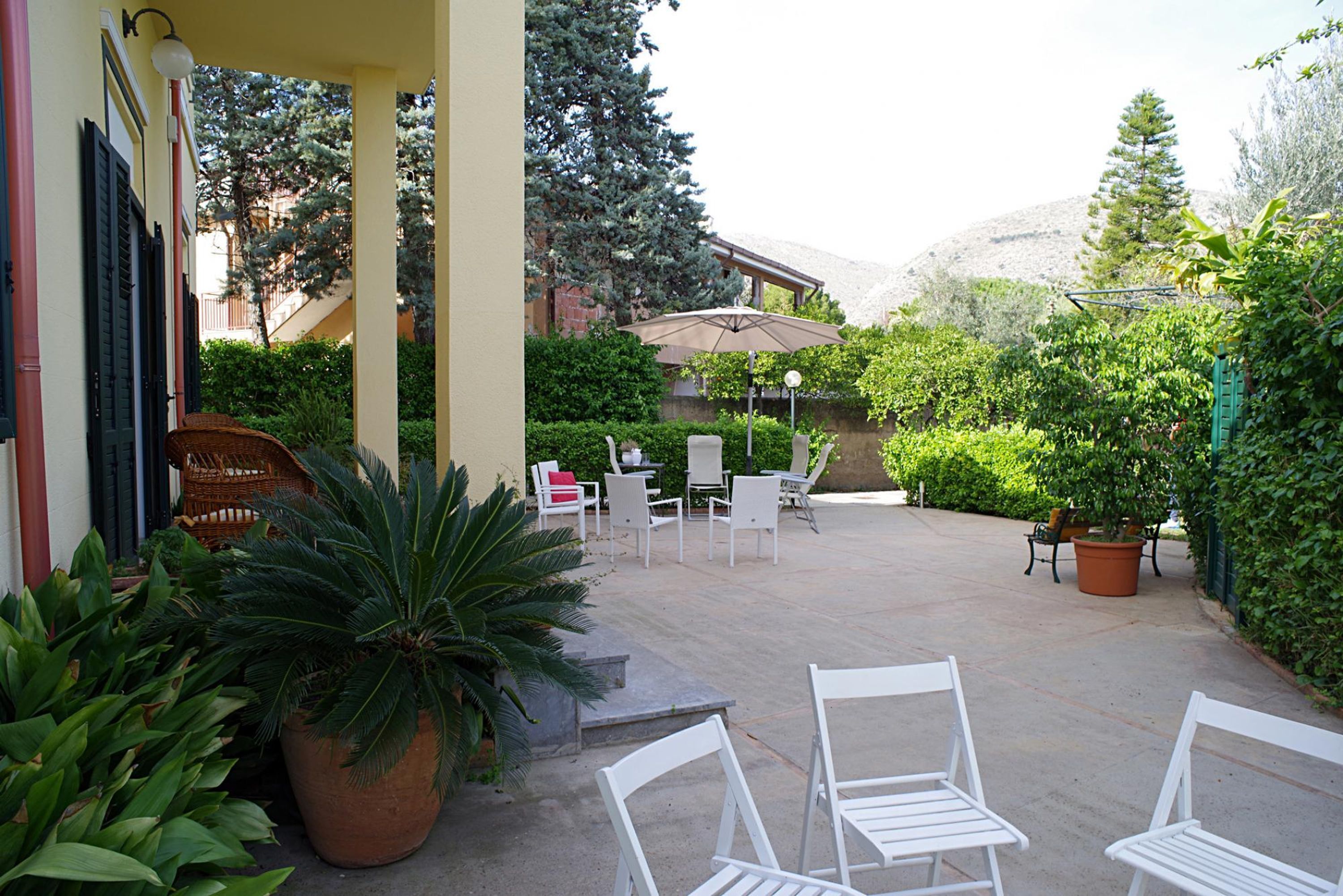 A covered outdoor seating area with white chairs and tables, surrounded by potted plants and greenery.