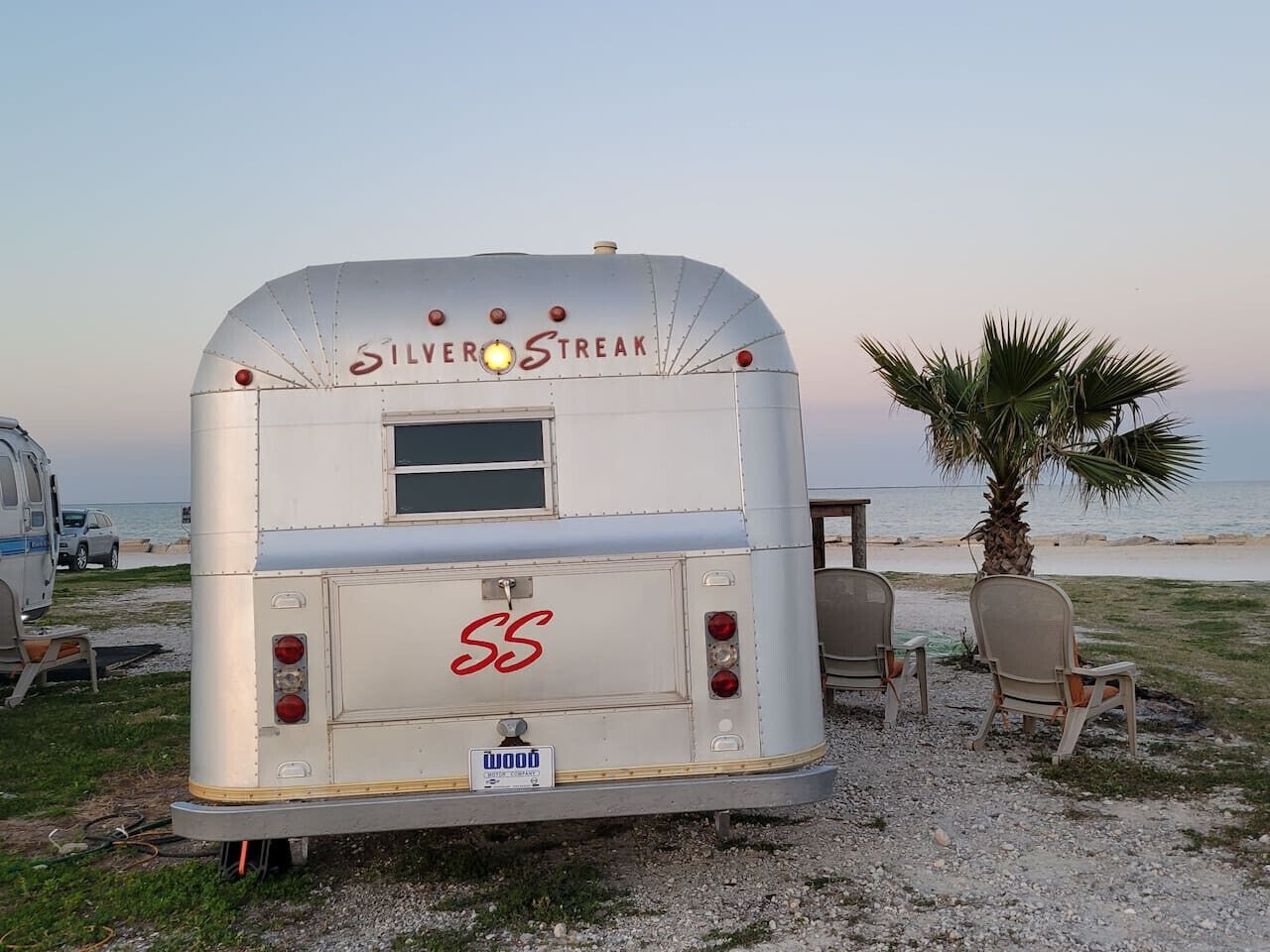 Een Silver Streak RV staat op het strand, met twee stoelen en een palmboom in de buurt.