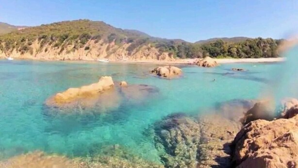 Paesaggio costiero con acque turchesi limpide, formazioni rocciose e uno sfondo collinare.