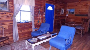 L’intérieur d’un chalet accueillant, avec des murs en bois, une porte bleue, deux fauteuils bleus, une petite table avec des magazines et une chaise en bois.