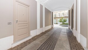 A corridor with a beige door, white walls, and a tiled floor leading to a courtyard with buildings and greenery. - Loft del Conte nei Pressi Della Mole by Wonderful Italy (Turin)