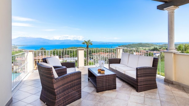 Ein Balkon mit Korbmöbeln, einem Glastisch und Blick auf Meer und Berge.
