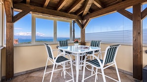 Ein überdachter Außenbereich mit Tisch und Stühlen, große Fenster mit Blick auf das Meer und die Berge sowie eine Holzbalkendecke.