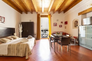 A bedroom with a bed, a desk, and a dining area. There is a window with curtains, a door, and a view of the outside.