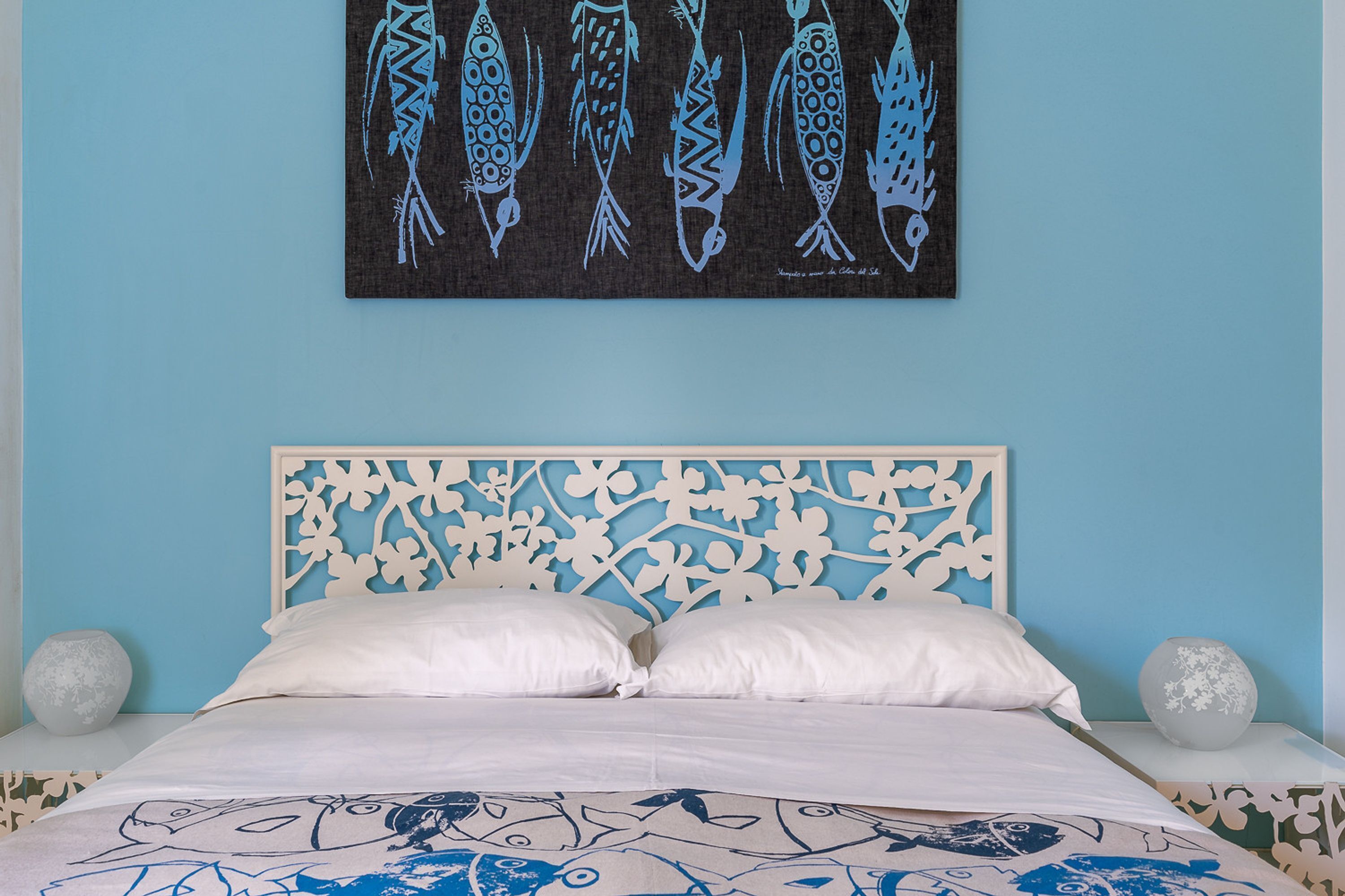 Een slaapkamer met een bed, witte kussens, een decoratief hoofdbord, een wanddecoratie van vissen en twee decoratieve vazen.