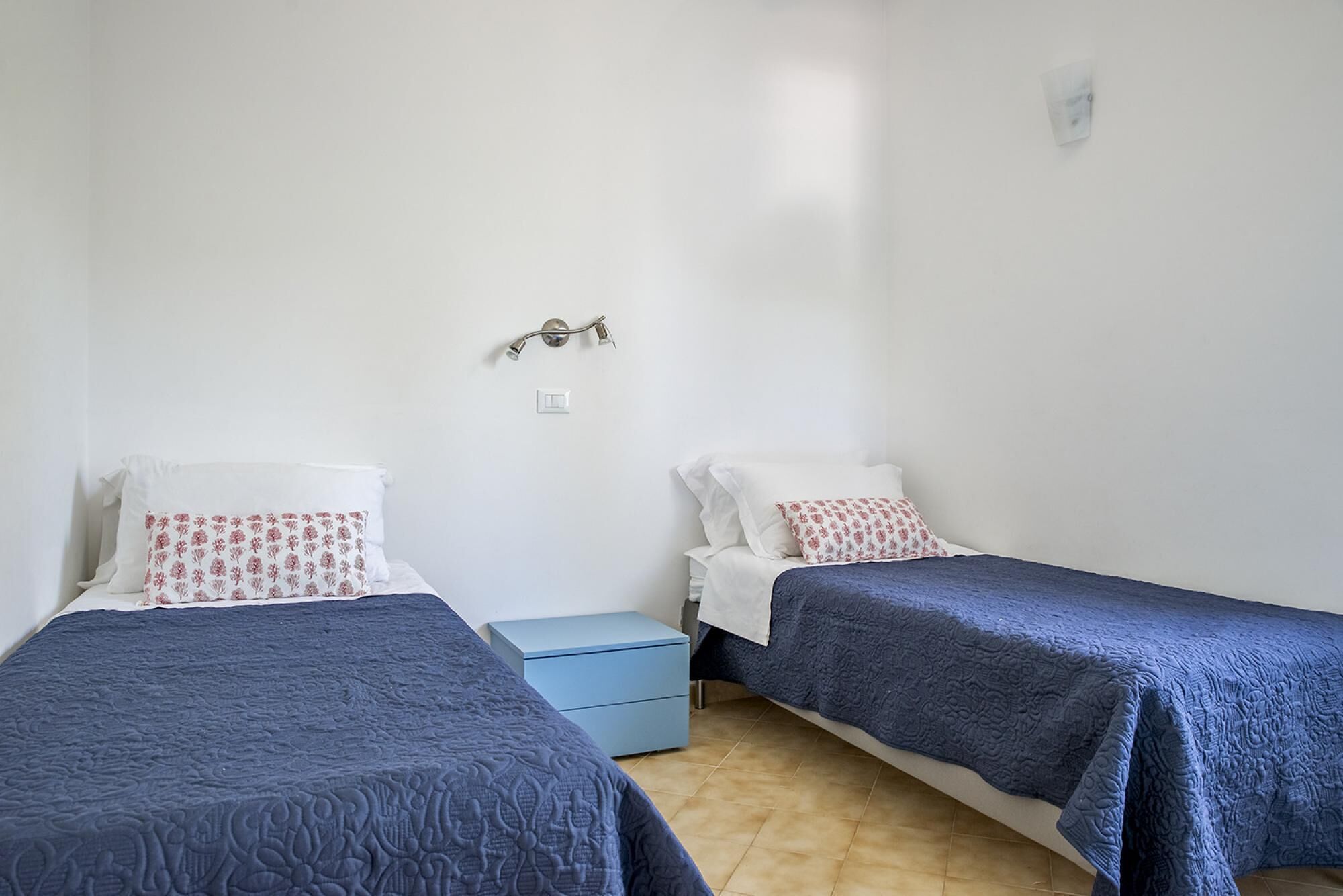 Quarto simples com duas camas, cada uma com colcha azul e almofadas brancas com estampas florais vermelhas. Há uma pequena mesa de cabeceira azul com abajur entre as camas.