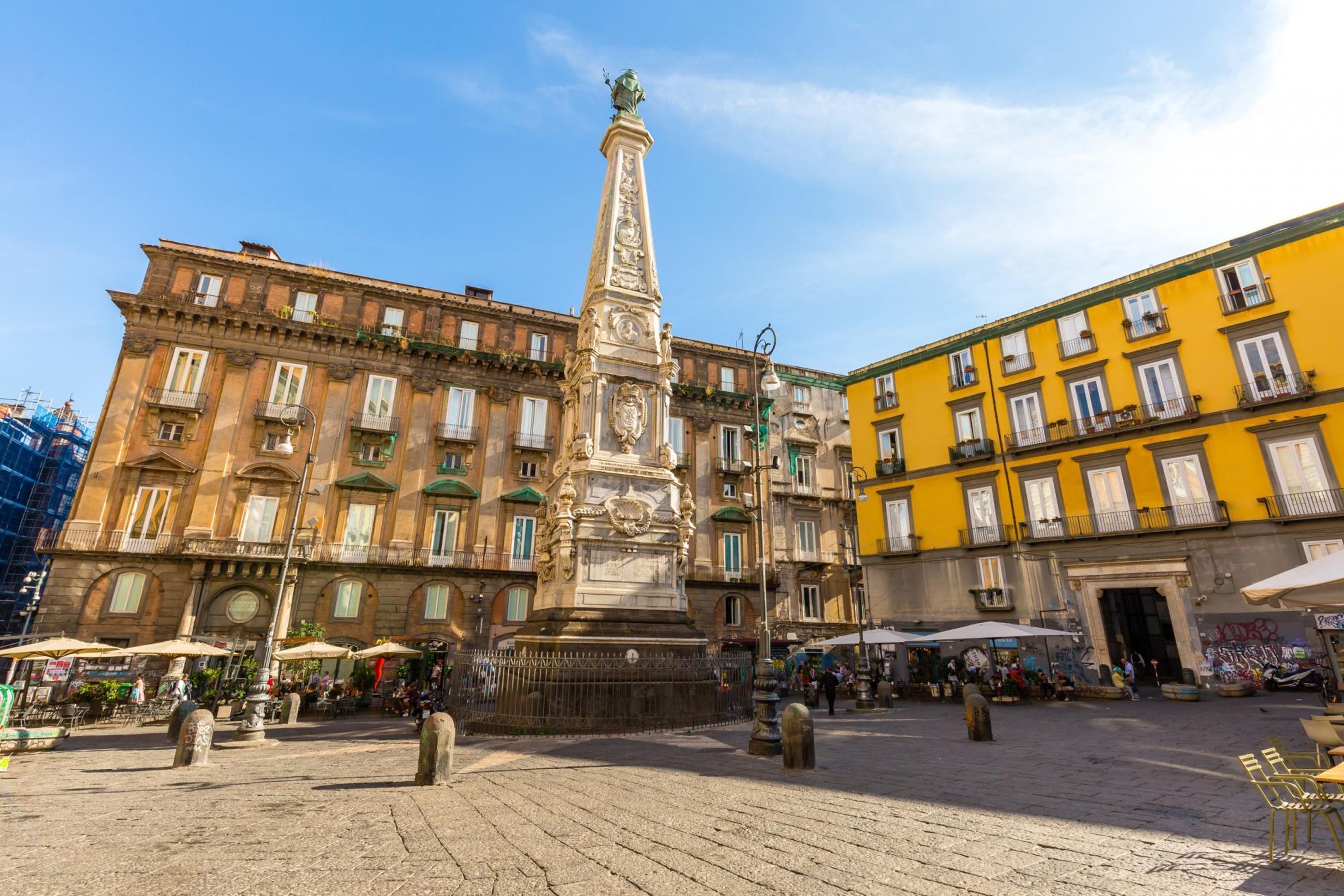 Ein Platz mit einem zentralen Denkmal, umgeben von historischen Gebäuden mit Balkonen und Außenbereichen mit Sitzgelegenheiten.