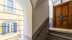 Un escalier menant à une porte en bois renforcée par du métal, une fenêtre avec des volets bleus et un mur muni d’un luminaire.