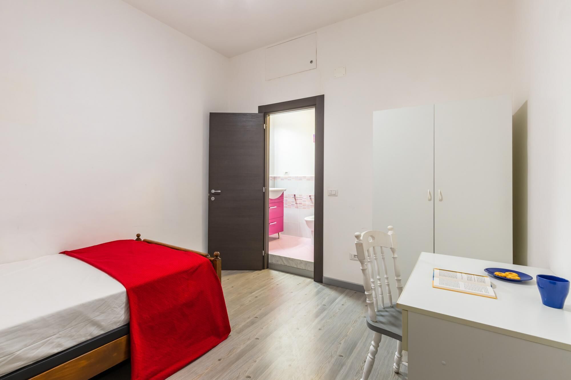 A small, well-lit room with a bed, a desk, and a wardrobe.