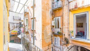 Une ruelle étroite en milieu urbain, bordée de bâtiments des deux côtés, avec des balcons ornés de plantes et de mobilier d’extérieur.