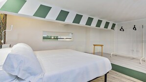 Quarto de hotel moderno com cama, uma mesinha, espelho e área de chuveiro.