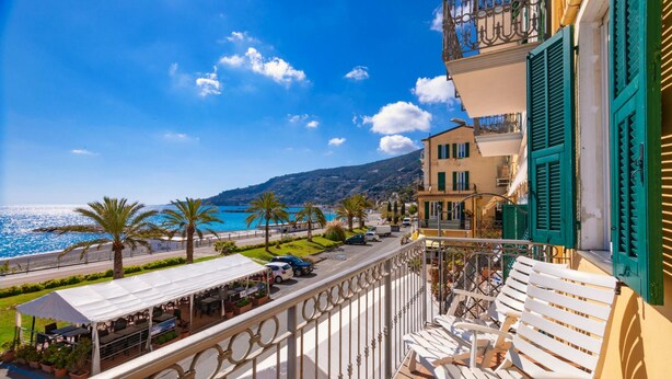 A balcony with white chairs, a view of the sea, and a building with green shutters.