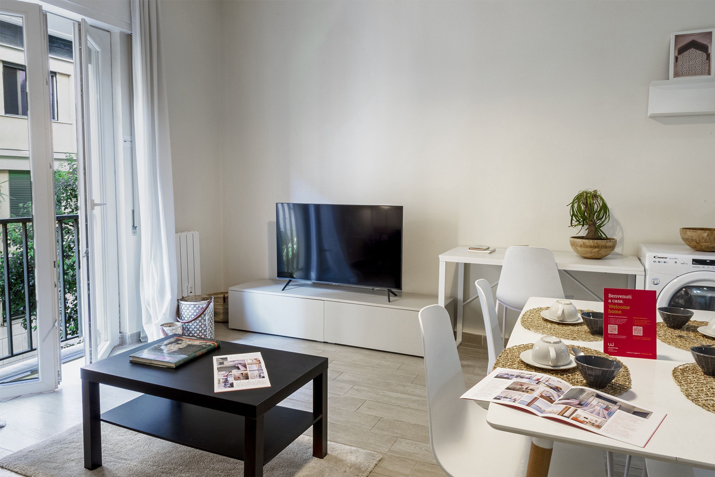 Een moderne woonkamer met een flatscreen televisie, een eettafel met een tijdschrift, een klein wit tafeltje en een rood welkom bord.