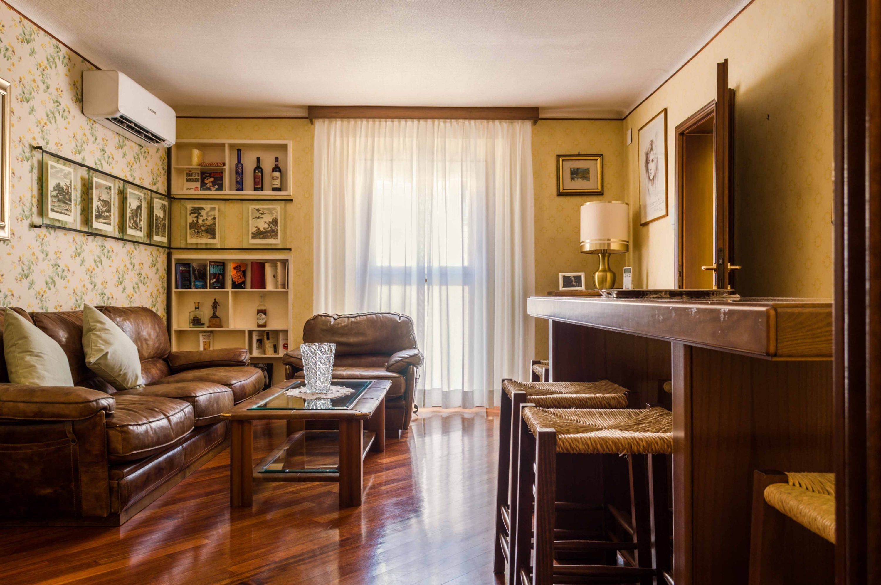 Een woonkamer met een bruine leren bank, een houten salontafel, rieten fauteuils, een boekenkast met ingekaderde foto’s en een open haard met een spiegel erboven.