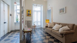 Um quarto com piso quadriculado, um sofá marrom com almofadas, uma pequena mesa de madeira, um abajur e uma janela com vista para o exterior.