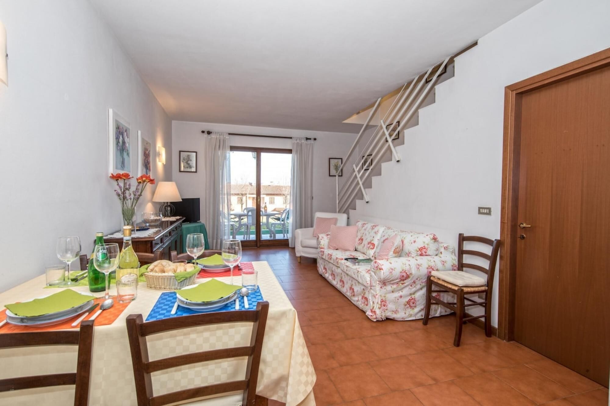 A dining area with a table set for two, a sofa, and a staircase leading to an upper level.