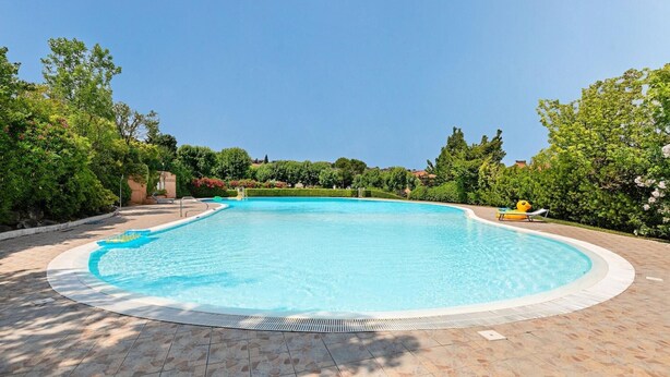 Une grande piscine aux eaux cristallines, bordée d’une terrasse carrelée et entourée d’une végétation luxuriante et d’arbres.