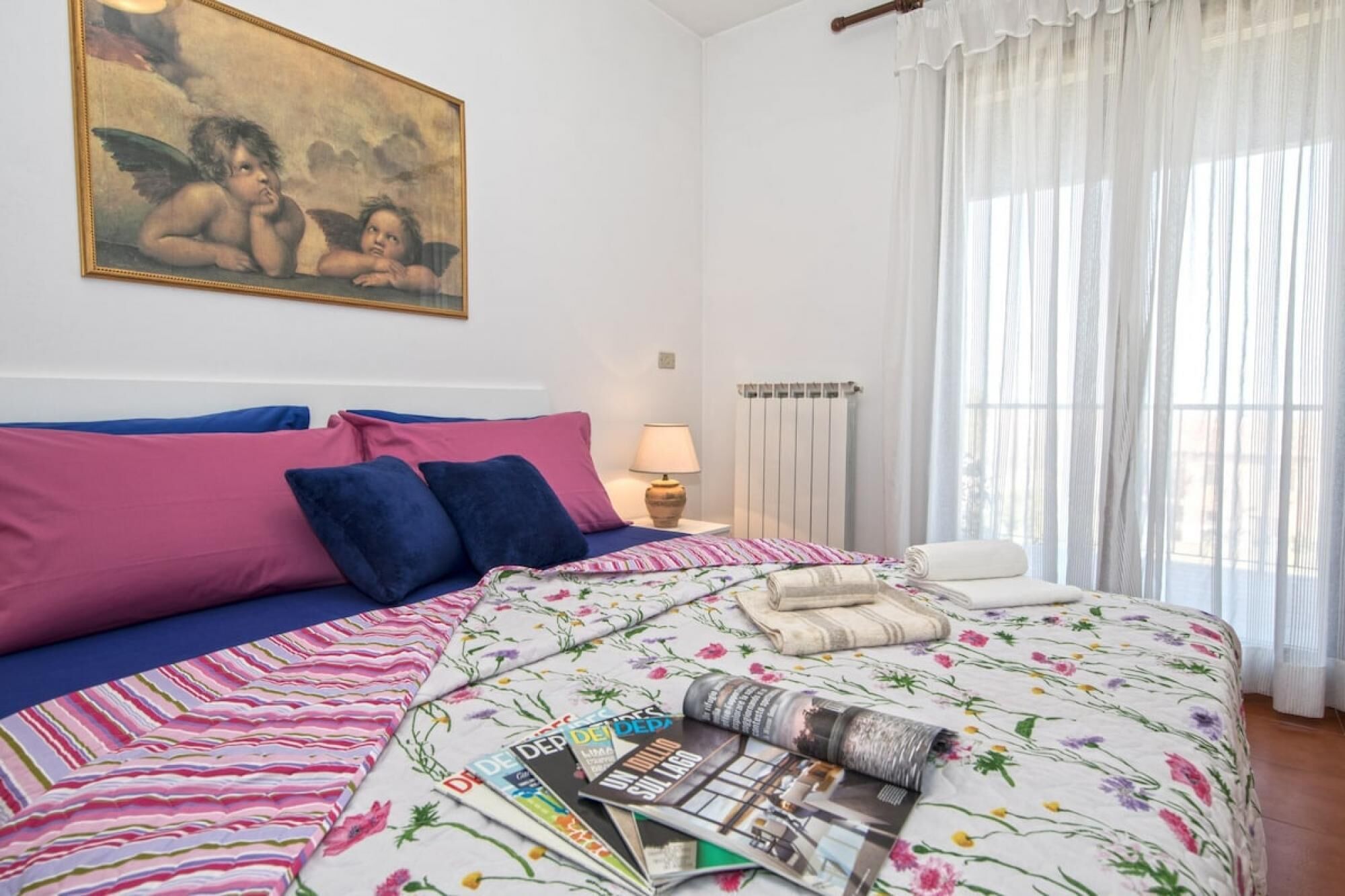 A bedroom with a bed, pillows, a lamp, a radiator, and a window with curtains.