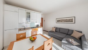 A modern kitchen with white cabinets and a dining table with a fruit bowl. A gray sofa and a framed picture are in the living area. - Residence Giardino 3 by Wonderful Italy (Soiano del Lago)