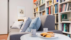 Una sala de estar moderna con un sofá gris, una mesa de centro blanca con pasteles, y una estantería llena de libros.