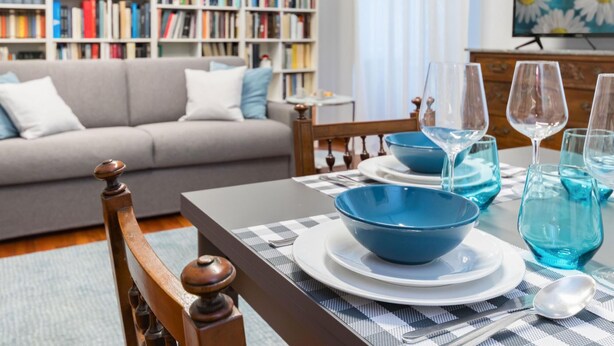Un comedor con una mesa dispuesta para cuatro personas, una estantería al fondo y un sofá en primer plano.