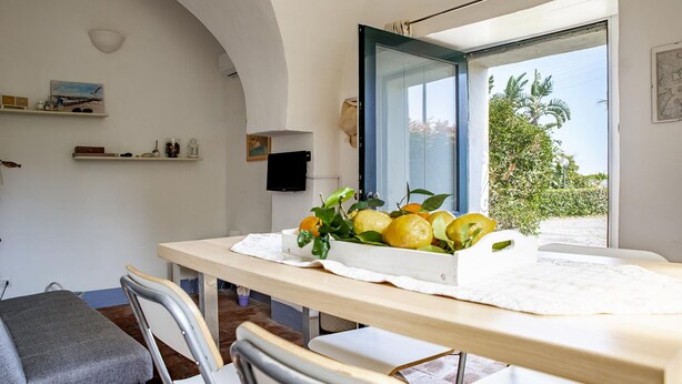 A dining area with a table set for two, a chair, a small shelf with decorative items, and a view of greenery outside. - Loft Sole by Wonderful Italy (Giarre)