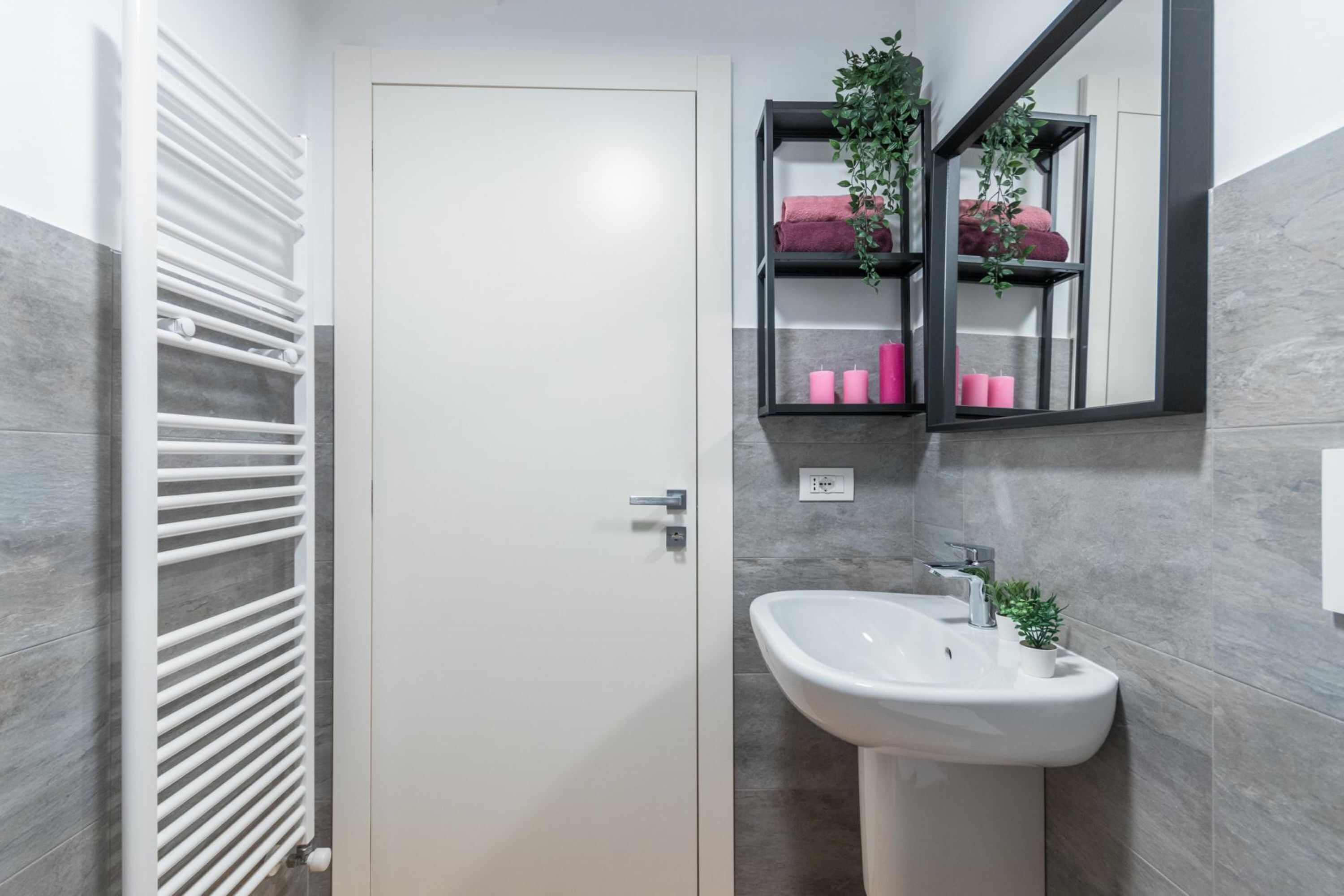A bathroom with a white door, a white radiator, a white sink, and a mirror with a shelf holding pink towels and candles.