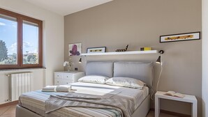 A bedroom with a bed, bedside tables, a nightstand, a window, and a wall-mounted shelf with decorative items. - Alessandra 2 Apartment by Wonderful Italy (Padenghe sul Garda)