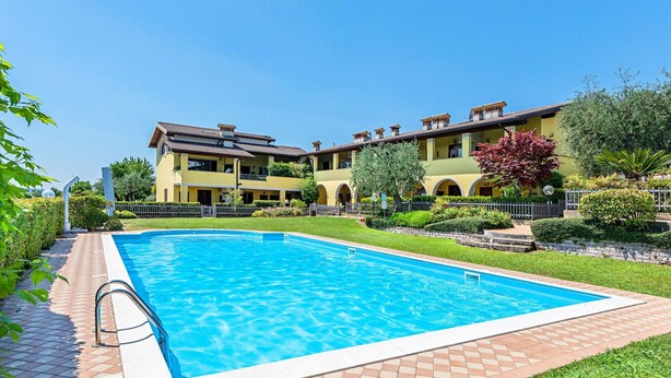 A swimming pool with a tiled deck, surrounded by a well-maintained garden and a multi-story building with balconies. - Alessandra 2 Apartment by Wonderful Italy (Padenghe sul Garda)