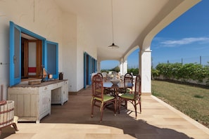 Un espace extérieur abrité, aménagé pour les repas, avec une table et des chaises, un buffet en bois et une vue sur la mer.